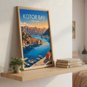 May include: A framed print of Kotor Bay, Montenegro, featuring a vibrant illustration of a coastal town with boats on the water and mountains in the background. The print is displayed on a wooden shelf with books and a small plant.