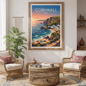 May include: A framed wall art print titled CORNWALL UK featuring a coastal landscape with cliffs, a sandy beach, a small village, and a sailboat on the water at sunset, displayed in a room with two wicker chairs and a round coffee table.