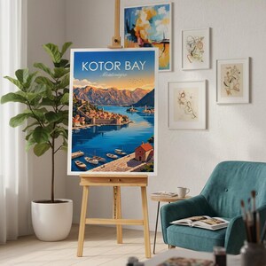 May include: A framed art print of Kotor Bay, Montenegro, displayed on a wooden easel. The print features a vibrant illustration of a coastal town with boats on the water, mountains, and the text "Kotor Bay Montenegro". The scene is set in a room with other framed art.