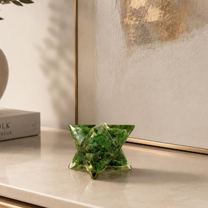 Può includere: Un oggetto decorativo a forma di stella realizzato in resina verde traslucida con texture incorporate simili a muschio verde. Il pezzo geometrico presenta molteplici vertici appuntiti e una finitura lucida che riflette la luce sulle superfici sfaccettate.