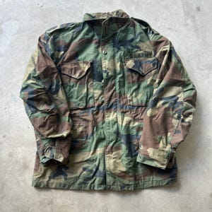May include: A long-sleeve field jacket featuring a woodland camouflage pattern in shades of green, brown, and black. The jacket includes a high collar, a front zipper closure with a snap-button placket, and two chest pockets with the text U.S. ARMY.