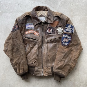 May include: Brown leather jacket with multiple patches, including an American flag, text that reads "MARTIAL ARTS", and a patch that reads "WEST BABYLON FIRE DEPARTMENT LONG ISLAND NEW YORK". The jacket has a zipper closure and ribbed cuffs.