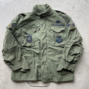 Op de afbeelding: Olijfgroene militaire jas van de US Air Force met meerdere patches en de tekst "U.S. AIR FORCE". De jas heeft een ritssluiting, meerdere zakken en een hoge kraag. Vintage kledingstuk.