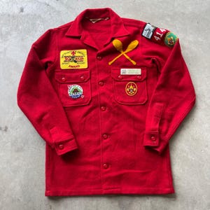 Op de afbeelding: Een rood shirt met lange mouwen in wolstijl met een knoopsluiting aan de voorzijde, twee borstzakken en diverse geborduurde patches, waaronder een Boy Scouts of America 50 Miler Award, een Frontier District 414 patch en twee gekruiste gele lepels.