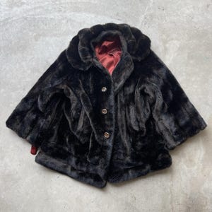 May include: A vintage black faux fur cape with a rounded collar and three button closure. The interior lining is a deep red color. The cape is a short length, and the fur has a soft, textured appearance. A classic outerwear piece.