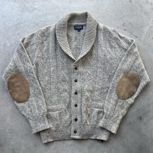Könnte beinhalten: Graue Strickjacke mit Zopfmuster, Schalkragen, Knopfverschluss und Ellbogenpatches aus Wildleder. Der Pullover hat zwei Vordertaschen und gerippte Bündchen. Das Lands' End-Etikett ist innen sichtbar.