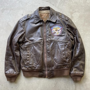 May include: A brown leather bomber jacket with a zip-up front, two front pockets, and a ribbed hem and cuffs. The jacket features a patch on the chest with text and illustrations. The jacket has a collared neckline and shoulder epaulets.