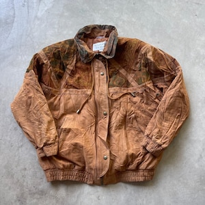 May include: A tan leather jacket featuring a textured brown patterned yoke across the shoulders and chest. The jacket includes a fold-down collar, a front zipper closure with a snap-button placket, long sleeves with elastic cuffs, and an elastic waistband.