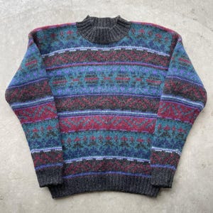 Puede incluir: Un suéter vintage con cuello alto y un colorido estampado geométrico. El suéter presenta rayas horizontales en tonos de azul, turquesa, morado y burdeos, con cuello y puños grises. El suéter está hecho de un material suave y tejido.