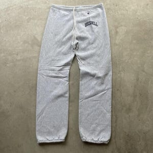 May include: Light grey Champion sweatpants with a drawstring waist and elasticated cuffs. The word "BUCKNELL" is arched across the upper left leg in navy blue. A small Champion logo is visible above the text.