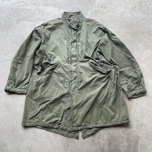 May include: An olive green military-style jacket with a button-up front, two large front pockets, and a stand-up collar. The jacket appears to be made of a lightweight material, with a slightly worn appearance. The jacket is a long coat.
