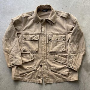 May include: A tan utility jacket featuring a collared neckline, long sleeves with buttoned cuffs, and four large front pockets with button closures. The jacket includes shoulder epaulets and a central button-down placket with a Polo by Ralph Lauren label.