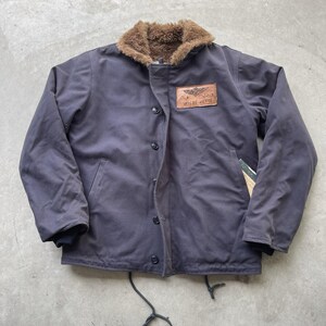 May include: A dark gray jacket featuring a brown faux fur collar, a front button closure, and a rectangular leather patch on the chest with a winged emblem and the text W.H. DE VETTE. The jacket includes long sleeves with ribbed cuffs and a drawstring hem.