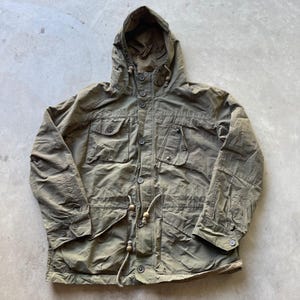 May include: An olive green hooded jacket featuring a front button closure, long sleeves with buttoned cuffs, and two chest pockets. The garment includes a drawstring waist with light-colored toggles and a hood with a drawstring adjustment.