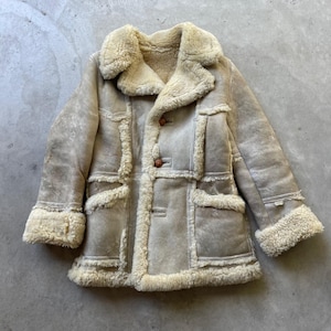 May include: A beige shearling coat featuring a wide collar, long sleeves with thick cuffs, and a front button closure. The garment is constructed with visible seams and exposed cream-colored fur trim along the edges, pockets, and hemline.