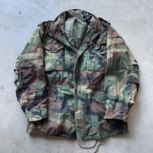 May include: A long-sleeve field jacket featuring a woodland camouflage pattern in shades of olive green, brown, and black. The jacket includes a high collar, a front zipper closure with a snap-button storm flap, and four large front pockets with flaps.