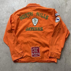 Puede incluir: Una chaqueta naranja de manga larga con texto bordado en verde que dice North Hills Jaycees. La parte posterior incluye un parche en forma de escudo, un parche circular y un parche rectangular rojo con texto blanco Jaycee State Board Meeting.