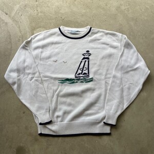 Könnte beinhalten: Ein weißer Langarmpullover mit Rundhalsausschnitt, der eine dunkelblaue Leuchtturm-Grafik mit grünen Wellen an der Basis und zwei kleinen Vögeln am Himmel zeigt. Der Pullover hat einen dunkelblauen Saum am Kragen, an den Ärmelbündchen und am unteren Rand.