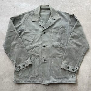 May include: A vintage olive green work jacket with a button-up front, two chest pockets, and two lower pockets. The jacket has the letters "USMC" stenciled on the chest pocket. The jacket has a worn appearance with frayed edges and some wear.