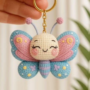 May include: A crochet butterfly keyring with a gold-coloured ring. The butterfly features a smiling face and is made in pink, blue, and cream. The wings have a polka dot and floral design.