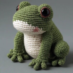 May include: A handmade crochet frog in shades of green and white. The frog has large, dark eyes with brown accents and a small pink blush on its cheek. The frog's body is a soft, textured material.