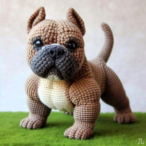 May include: A crocheted brown and tan dog figurine with a distinctive bulldog-like face. The handmade plush toy has a textured appearance, with a gray muzzle and black eyes. The dog is posed on a patch of green, resembling grass.