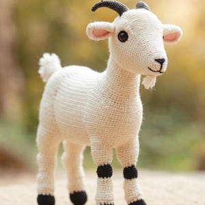 May include: A crocheted goat toy, crafted from off-white yarn. It has black horns, hooves, and knee markings. The goat has a small white beard, pink inner ears, and brown eyes. It stands on a light brown surface.