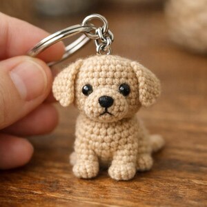 May include: A crocheted puppy keychain in a light tan color. The small dog has black eyes and a black nose. The keychain is attached to a silver metal ring. The puppy is sitting on a wooden surface.