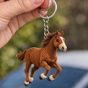 May include: A brown crocheted horse keychain with a white blaze on its face and white socks on its hooves. The horse is in a running pose, attached to a silver keyring and chain.