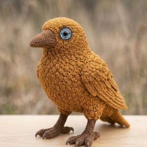 May include: A handcrafted, crochet bird sculpture in shades of brown and tan. The bird has detailed wings, feet, and a beak. It features large, striking blue eyes, adding a whimsical touch to the handmade creation. A unique decorative item.