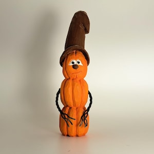 May include: A hand-carved wooden figurine shaped like a stacked orange pumpkin character wearing a tall brown hat. The figure features painted white eyes with black pupils, a small brown nose, and thin black wire arms extending from the sides.