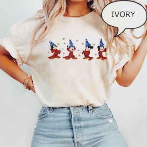 K&ouml;nnte beinhalten: Elfenbeinfarbenes T-Shirt mit einer Grafik von vier Zeichentrickfiguren in roten Roben und blauen Zaubererh&uuml;ten. Die Figuren sind in verschiedenen Posen mit goldenen Sternen. Das Wort "IVORY" befindet sich in einer Sprechblase.