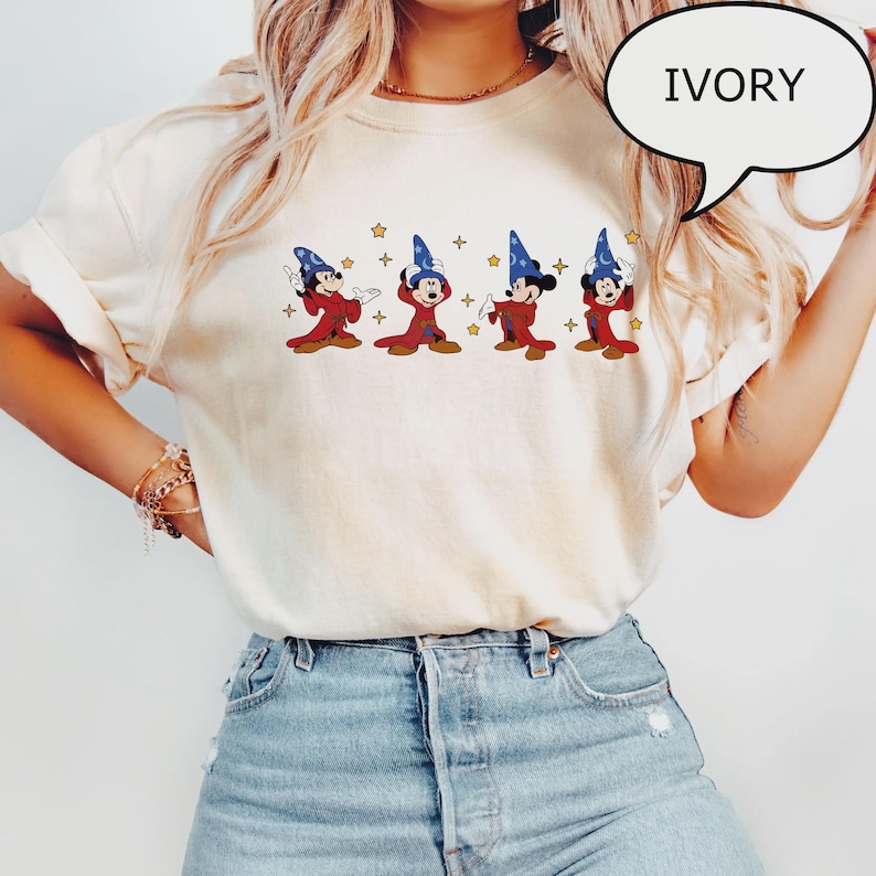 K&ouml;nnte beinhalten: Elfenbeinfarbenes T-Shirt mit einer Grafik von vier Zeichentrickfiguren in roten Roben und blauen Zaubererh&uuml;ten. Die Figuren sind in verschiedenen Posen mit goldenen Sternen. Das Wort "IVORY" befindet sich in einer Sprechblase.