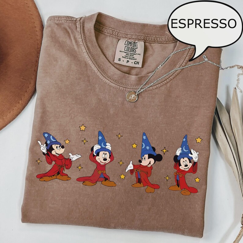 K&ouml;nnte beinhalten: Espressofarbenes T-Shirt mit einem Aufdruck von Mickey Mouse in einem Zauberer-Outfit. Das Design zeigt Mickey in einem roten Gewand und einem blauen Zaubererhut mit gelben Sternen. Das Shirt hat ein Comfort Colors-Etikett und eine silberne Halskette.