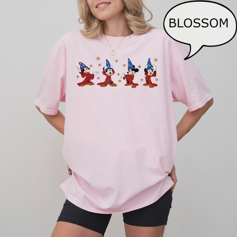 K&ouml;nnte beinhalten: Hellrosa T-Shirt mit einem Cartoon-Design von Mickey Mouse als Zauberer. Das Design zeigt vier Mickey Mouse Figuren in roten Roben und blauen Zauberh&uuml;ten, mit goldenen Sternen. Das Wort "BLOSSOM" befindet sich in einer Sprechblase.