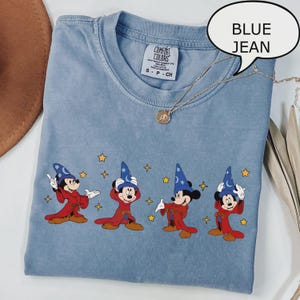 K&ouml;nnte beinhalten: Hellblaues T-Shirt mit einem Aufdruck von Mickey Mouse in einem Zauberkost&uuml;m. Das Shirt hat einen Rundhalsausschnitt und kurze &Auml;rmel. Eine goldene Halskette mit Anh&auml;nger ist &uuml;ber dem Shirt drapiert.
