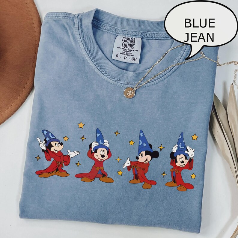 K&ouml;nnte beinhalten: Hellblaues T-Shirt mit einem Aufdruck von Mickey Mouse in einem Zauberkost&uuml;m. Das Shirt hat einen Rundhalsausschnitt und kurze &Auml;rmel. Eine goldene Halskette mit Anh&auml;nger ist &uuml;ber dem Shirt drapiert.