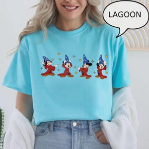 K&ouml;nnte beinhalten: Hellblaues T-Shirt mit einer Grafik von Mickey Mouse in einem Zauberer-Outfit. Das Design zeigt Mickey in verschiedenen Posen mit rotem Gewand, blauem Hut und goldenen Sternen. Das Wort "LAGOON" steht in einer Sprechblase.