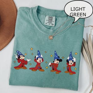 K&ouml;nnte beinhalten: Hellgr&uuml;nes T-Shirt mit einer Grafik von Mickey Mouse in einem Zauberer-Outfit. Das Design zeigt Mickey in verschiedenen Posen mit einem roten Umhang und einem blauen Zaubererhut. Das T-Shirt hat einen Rundhalsausschnitt und kurze &Auml;rmel. Eine silberne Halskette ist &uuml;ber dem T-Shirt drapiert.