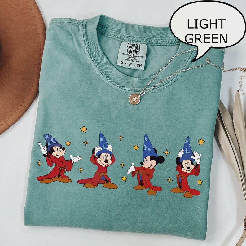 K&ouml;nnte beinhalten: Hellgr&uuml;nes T-Shirt mit einer Grafik von Mickey Mouse in einem Zauberer-Outfit. Das Design zeigt Mickey in verschiedenen Posen mit einem roten Umhang und einem blauen Zaubererhut. Das T-Shirt hat einen Rundhalsausschnitt und kurze &Auml;rmel. Eine silberne Halskette ist &uuml;ber dem T-Shirt drapiert.
