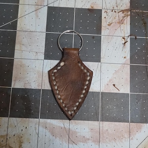 May include: A brown leather keychain with a shield shape and a silver key ring. The edges are stitched with white thread. The keychain is on a gray and black checkered surface.