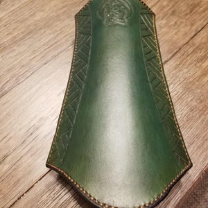 Puede incluir: Un protector de brazo de cuero verde con un diseño de nudo celta en la parte superior. El protector de brazo tiene costuras decorativas y una forma curva. El cuero tiene un acabado liso y un color verde oscuro.