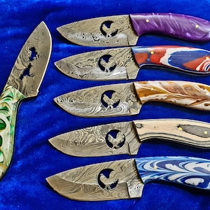 Puede incluir: Un surtido de cuchillos hechos a mano con hojas estampadas y mangos coloridos. Las hojas presentan un diseño recortado de gallo. Los colores de los mangos incluyen púrpura, rojo, blanco, azul, marrón y verde. Los cuchillos se exhiben sobre una superficie azul.