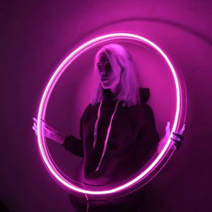 May include: A person is framed by a glowing pink neon circle. The person is wearing a dark hoodie. The background is a dark purple colour. The neon circle is held in front of the person.