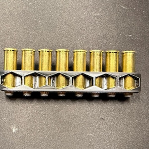 Può includere: Un supporto per munizioni in plastica nera con una griglia a nido d'ape contenente otto cartucce metalliche color ottone. Il supporto è rettangolare e presenta fessure esagonali individuali per ogni cartuccia, disposte in una fila.