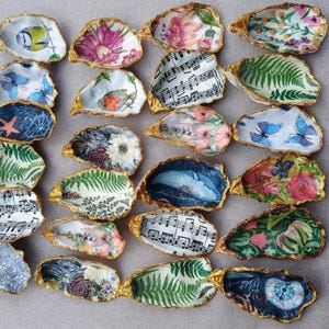 May include: A collection of twenty-four decorative oyster shells, each featuring a unique decoupage design including floral patterns, butterflies, musical notes, ferns, birds, and marine life, all finished with a gold-painted scalloped edge.