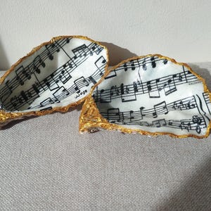 Peut inclure: Deux plats décoratifs en forme de coquilles d'huîtres. L'intérieur de chaque coquille est blanc avec des notes de musique noires. Les bords sont garnis de peinture dorée. Les coquilles reposent sur une surface grise.
