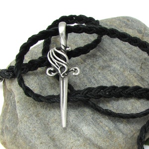 May include: A silver pendant in the shape of a stylized dagger with a black braided cord necklace. The pendant is on a gray rock.