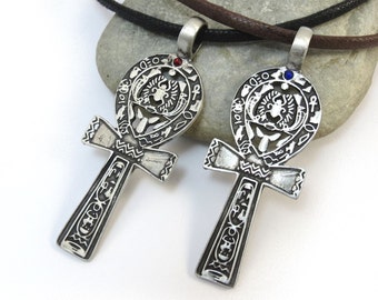 May include: Two silver Ankh pendants with intricate designs and a red and blue gemstone in the center of each. The pendants are on a brown and black cord.