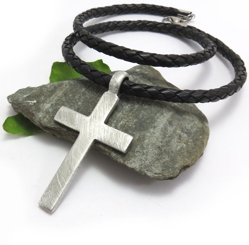 May include: A silver cross pendant on a black braided leather cord necklace. The cross has a brushed finish and is hanging on a rock.